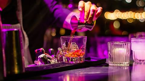 Bartender pouring a cocktail into a glass at a well-lit bar counter.