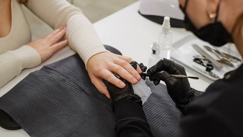 A manicurist wearing black gloves applies nail polish to a client's nails at a salon table with nail care tools on it.
