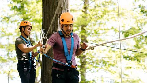 Two people wearing helmets and harnesses participate in a rope course among trees.