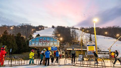 Skiers gather at the base of a snowy slope at dusk, preparing to ascend the mountain. The Coors Light logo is visible on the lift station.
