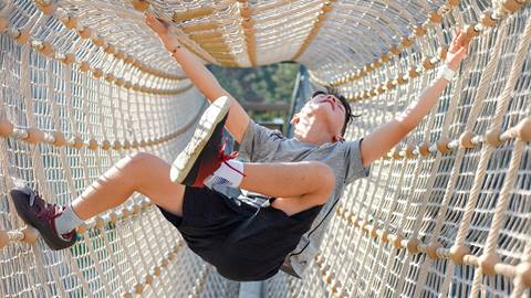 A person in athletic wear climbs inside a rope tunnel bridge, balancing with arms and legs extended.