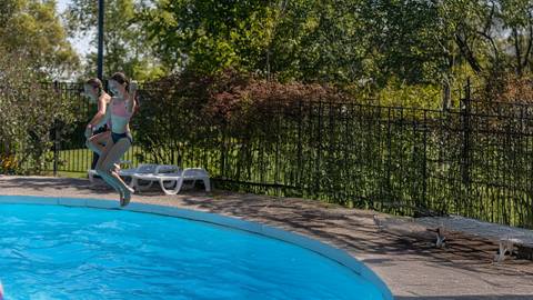 Two girls in swimsuits and goggles are jumping into an outdoor swimming pool on a sunny day, with trees and lounge chairs in the background.