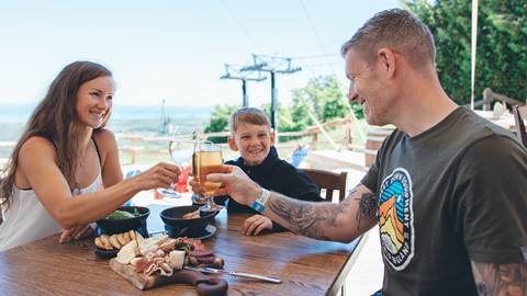 A woman, man, and child clink glasses while sitting at an outdoor table with food on a sunny day near ski lift structures.