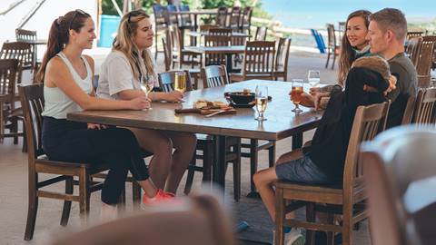 Five people sit around a wooden table outdoors, eating, drinking, and talking. Plates, glasses, and food are visible on the table. Empty chairs and sea are in the background.