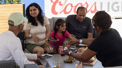 Five people sit around a table outdoors, sharing food and drinks, with a large "Coors Light" sign visible in the background.