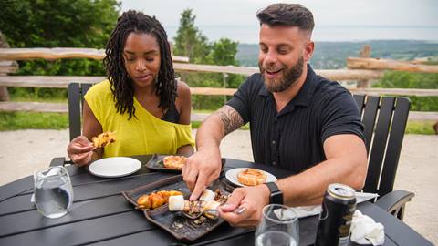 Two people sit at an outdoor table, sharing a meal with plates of food and drinks in front of them, with a scenic landscape in the background.