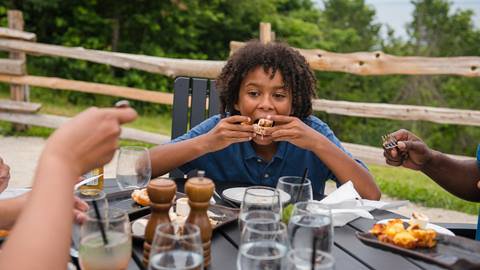 A child sits at an outdoor table, eating a sandwich with both hands while others dine around him; plates and glasses are visible on the table.
