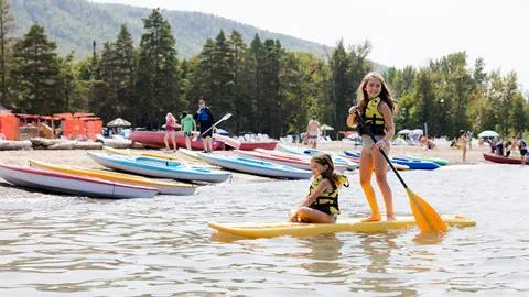 Two girls with life jackets on a paddleboard at a lake shore, with kayaks in the background.