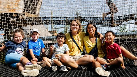 A group of children and two adults sitting on a net at a playground, smiling for the camera.