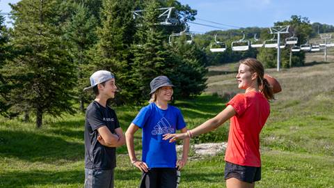 Three young people enjoying a sunny day outdoors with a frisbee, with a chairlift and green hill in the background.