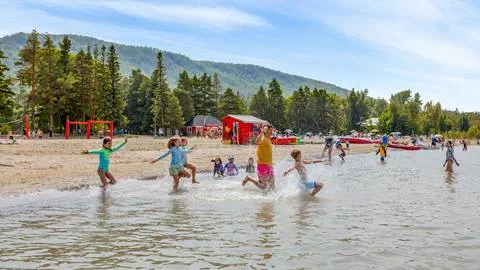 Children and adults playing and splashing in the shallow waters of a beach with a forested hill in the background.