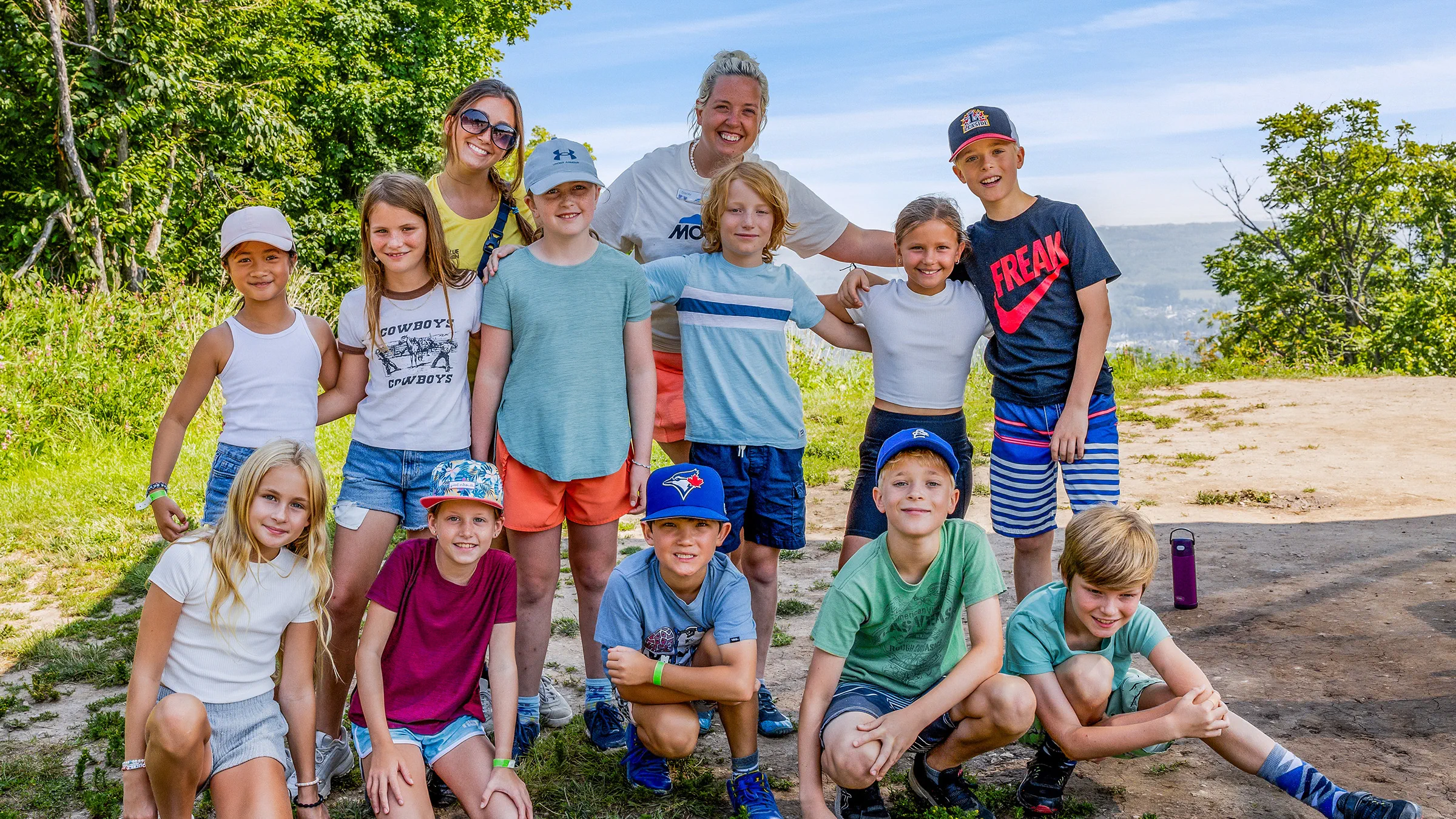 Group of children with two adults posing for a photo on a sunny day with a scenic overlook in the background.