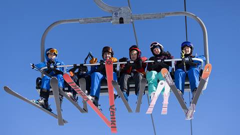Skiers wearing helmets and goggles seated on a chairlift with their skis dangling below.