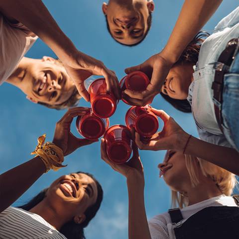 Five people stand in a circle outdoors holding red plastic cups together, viewed from below with the blue sky in the background.