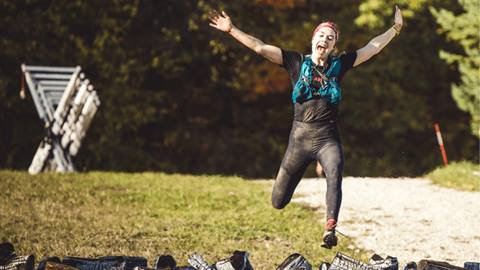 A person with arms raised jumps over a row of black rocks or obstacles outdoors, wearing athletic gear and a hydration pack. Trees and grass are visible in the background.