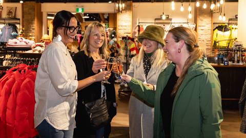 Four women standing in a clothing store clink glasses together, smiling and appearing to celebrate. Coats and jackets are displayed in the background.