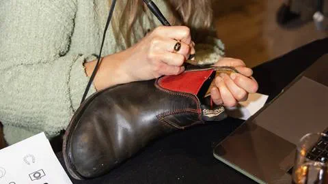 A person uses a tool to repair or customize a black Blundstone boot with red lining, holding it steady near a laptop on a table.