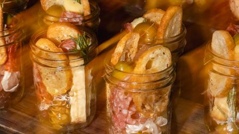 Glass jars filled with crackers, cheese, olives, sliced meats, and rosemary sprigs arranged on a wooden surface.