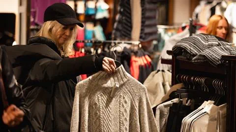 A woman in a black jacket and cap examines a gray sweater in a clothing store, surrounded by racks of various garments.