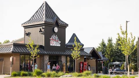 A group of people stands outside a building with a clock tower and a sign reading "Red Devil Sports." There are trees and a vending machine nearby.