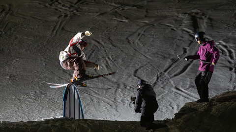 A skier performs a trick on a rail at night while two people film the action on a snowy slope.