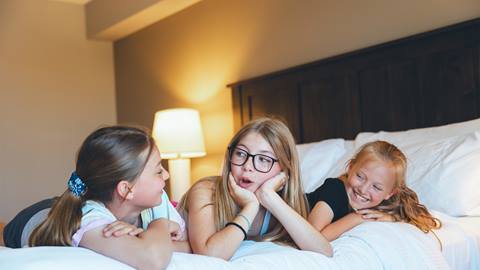 Three girls lying on a bed, talking and smiling, with a bedside lamp and headboard visible in the background.