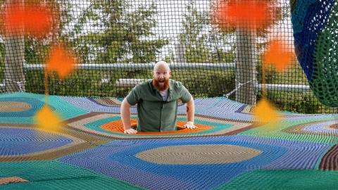 A man with a beard and green shirt sits partially inside a colorful net structure outdoors, surrounded by trees and netting.