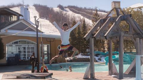 A boy in swim trunks jumps into an outdoor pool with snow-covered hills in the background; other people are in and around the pool.